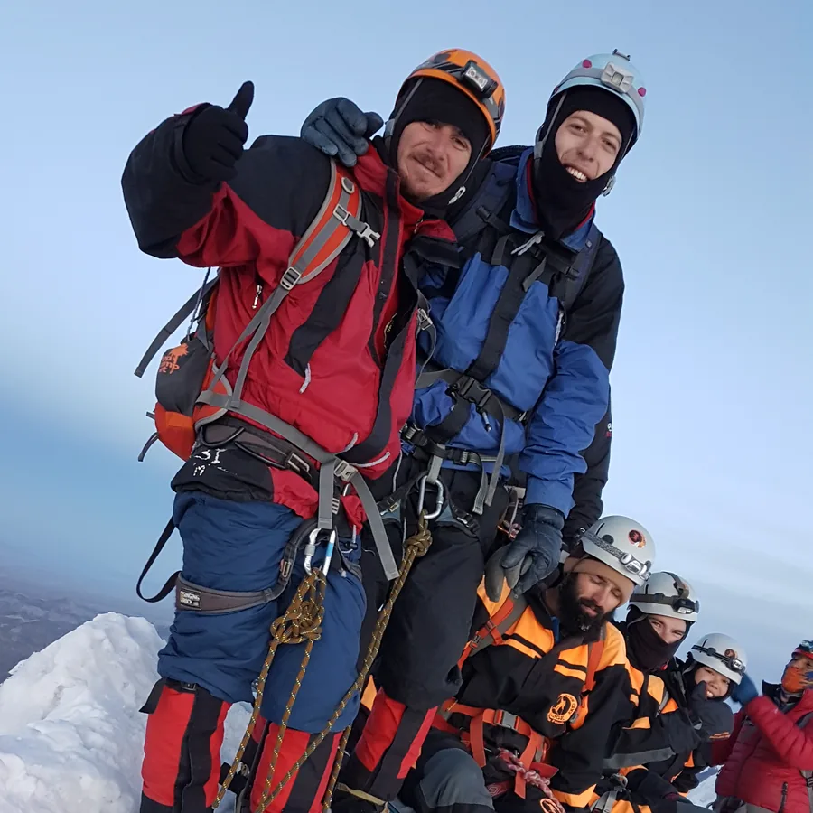 A roped mountaineering team of six on a narrow snow summit above the cloud line, in helmets, harnesses and crampons, ropes running between them.