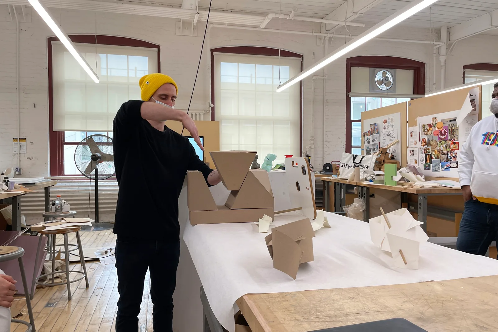 Tomer standing at a studio crit table in a Pratt Institute workshop, leaning on a stack of cardboard form models, with project boards and other students' work behind him.
