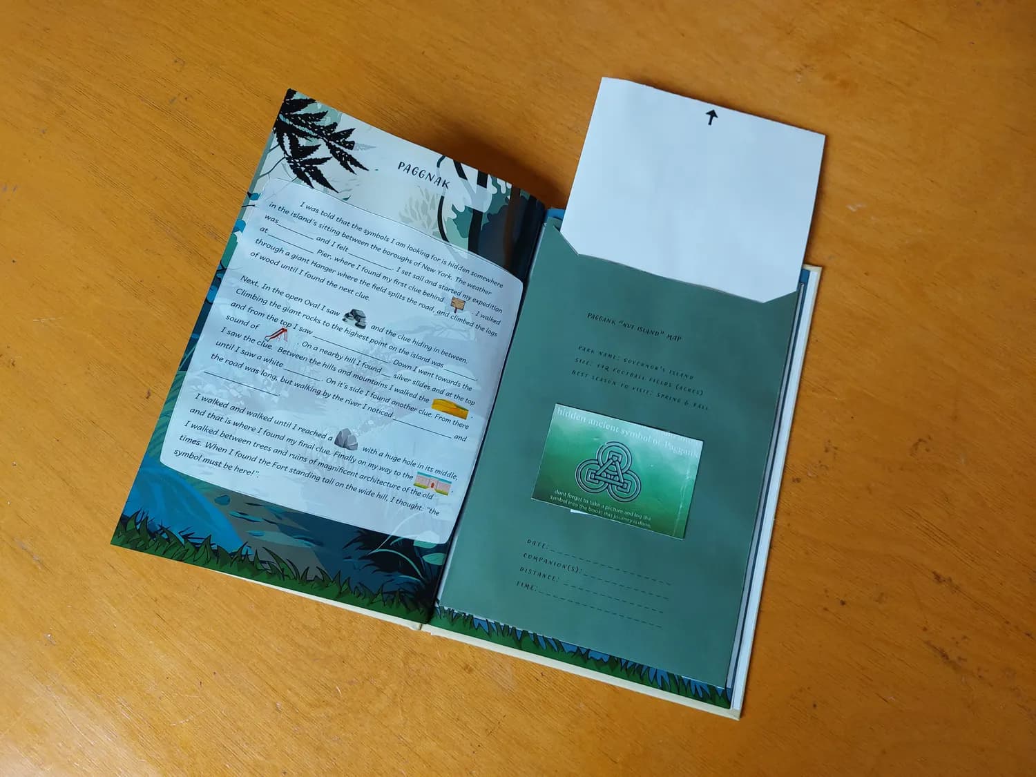 The activity book open on a table, showing an illustrated narrative page on the left and a pocket on the right holding the fold-out park map and a collectible card.